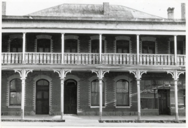 Black and white photograph of two storey brick hotel with corrugated iron roof and three chimneys. Second storey shows seven doors leading to balcony with ornamental railing, supported by five columns. Ground floor shows a window, two doors, two windows, and a storefront.