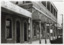 Black and white photograph taken down footpath at an angle, shows awning of R. Rosen tobacconist and two storey brick hotel next door. Hotel balcony rail supported by seven columns present, ground floor shows two windows separated by two doors, and a storefront. Buildings continue in distance, front left of car in bottom right of photo.