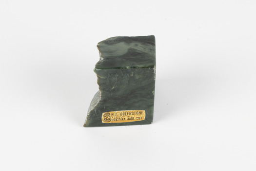 Solid mineral specimen of smooth grey-green rock with pale veins and mottling, marked with a gold sticker that reads "N.Z. Greenstone/ a product of/ Hokitika Jade Coy."