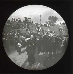 A monochrome photograph within a circle frame featuring a group of men, women and children dressed in 19th century clothing standing in front of, and on, a small rocky hill. 