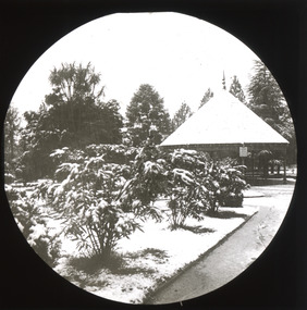 A monochrome photograph within a circle frame featuring a snow-covered avenue of shrubs leading to an open wooden rotunda with a conical roof within a large garden.