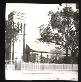 A monochrome photograph within a square frame featuring an image of a single-storey stone church with a taller square tower and narrow arched windows, set in a garden surrounded by a low picket fence. 