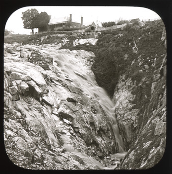 A monochrome photograph within a round-cornered square frame depicting buildings at the top of the frame above a rocky incline and small waterfall.