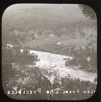 A monochrome photograph within a round-cornered square frame showing properties and pastoral land bordering a river, taken from a height.