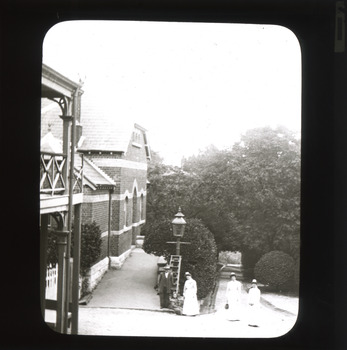 A monochrome photograph within a rectangular frame showing three women and one man in early Twentieth-Century attire near a large brick building with gardens behind them. 