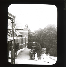 A monochrome photograph within a rectangular frame showing three women and one man in early Twentieth-Century attire near a large brick building with gardens behind them. 