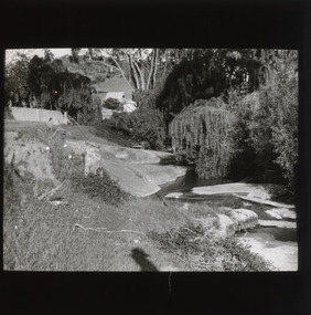 A monochrome photograph within a rectangular frame showing properties and shrubbery bordering a river or stream flowing downhill over rocks.