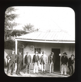 A monochrome photograph within a round-edged square frame showing eight men standing in a row outside a small tin-roofed wooden building.