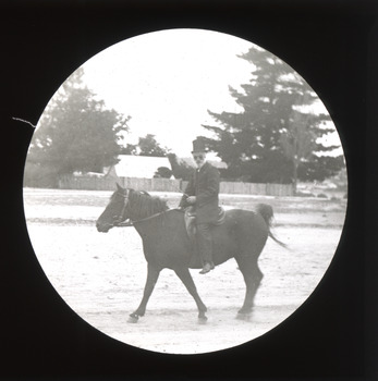 A monochrome photograph displaying a round image of a man in a top-hat and suit riding a horse along a snow-covered roadway with houses in the background. 