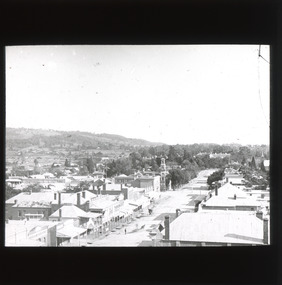 A monochrome photograph with a rectangular frame showing a view of shops and other buildings along a major thoroughfare in a country town, taken from a height.