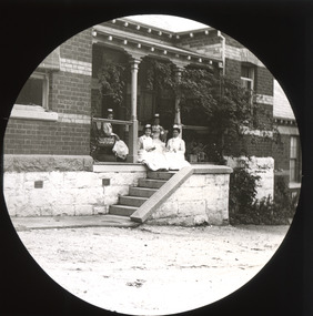 A monochrome photograph within a circular frame showing four women in early Twentieth-Century nurses' uniforms seated on a bench and steps at the entry to a large building. 