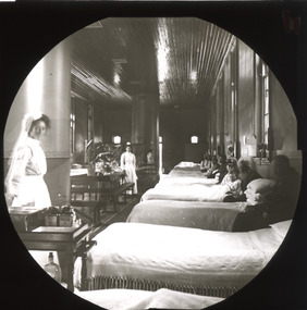 A monochrome photograph within a circle frame featuring a large hospital ward with a receding row of beds occupied by men and boys and three standing women dressed in early twentieth century nursing uniforms.  