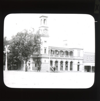 A monochrome photograph within a round-edged square frame featuring a 19th century styled double-storied building with a tall clock tower on its left side and a colonnaded entrance. 