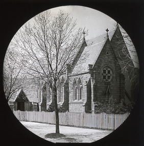 A monochrome photograph within a circle frame featuring an image of a fenced-in brick building with a triangular slanting roof and a row of arched windows. A leafless tree stands in the forefront of the image, adjacent to the building.  