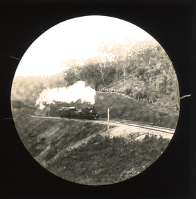 A monochrome photograph within a circular frame featuring an image of a steam train proceeding along a hillside rail track in a rural setting. 
