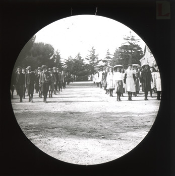 A monochrome photograph within a circular frame depicting older children wearing the clothing of the late 19th/early 20th Centuries walking in gender-divided lines towards the camera in an open area adjacent to a school building.
