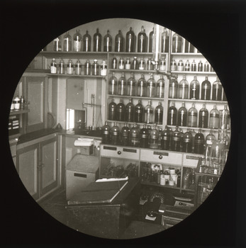 A monochrome photograph within a circular frame depicting tightly-packed floor-to-ceiling shelving filled with transparent storage and dispensing bottles. Writing implements and pharmacy equipment of the early Twentieth Century is also visible. 