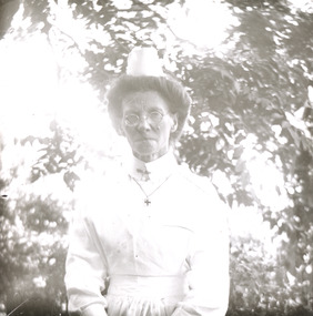 A monochrome photograph featuring a portrait image of an older woman from the waist up dressed in an early twentieth century styled white nurse's uniform. 