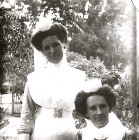 A monochrome photograph featuring a portrait image of two women - one standing and one sitting - dressed in early-twentieth century styled white nurses uniforms. 