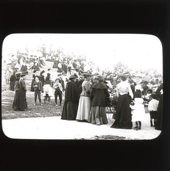 A monochromatic photograph within a rectangular frame with curved edges featuring a group of women, men and children dressed in 19th century clothing and standing in a park and atop a large rock.