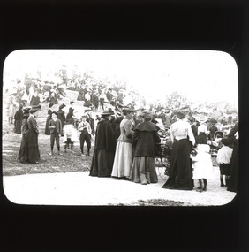 A monochromatic photograph within a rectangular frame with curved edges featuring a group of women, men and children dressed in 19th century clothing and standing in a park and atop a large rock.