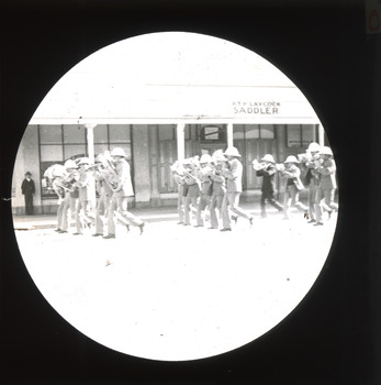 A monochromatic photograph within a circular frame featuring three rows, five people in each, wearing marching band uniforms with instruments marching down a street in front of RTP LAYCOCK SADDLER