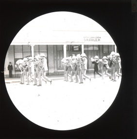 A monochromatic photograph within a circular frame featuring three rows, five people in each, wearing marching band uniforms with instruments marching down a street in front of RTP LAYCOCK SADDLER