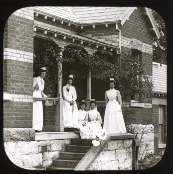 A monochrome photograph within a square frame featuring an image of five nurses gathered on a porch and staircase.