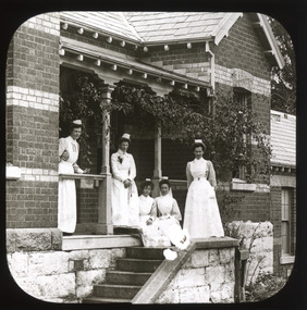 A monochrome photograph within a square frame featuring an image of five nurses gathered on a porch and staircase.