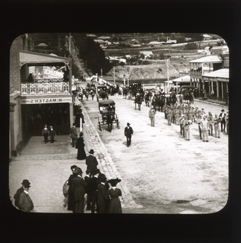 A monochrome photograph within a square frame featuring an image of military parade down a street watched by onlookers gathered on the sidewalks.