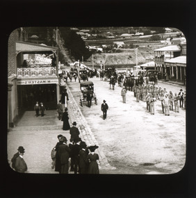 A monochrome photograph within a square frame featuring an image of military parade down a street watched by onlookers gathered on the sidewalks.