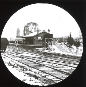 A monochrome photograph within a circular frame featuring an image of a rural railway station, showing multiply train tracks and the small station building with attached platform.