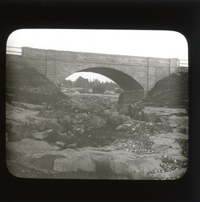 A monochrome photograph within a square frame featuring an image of stone bridge across a gorge.