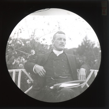 A monochrome photograph within a circular frame featuring an image of a well-dressed man casual sitting on a bench seat outdoors with a pile of papers balanced on his crossed legs.