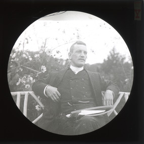 A monochrome photograph within a circular frame featuring an image of a well-dressed man casual sitting on a bench seat outdoors with a pile of papers balanced on his crossed legs.