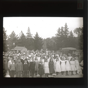 A monochrome photograph within a circle frame featuring a group of school children standing in front of some tall trees.