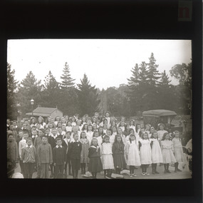 A monochrome photograph within a circle frame featuring a group of school children standing in front of some tall trees.