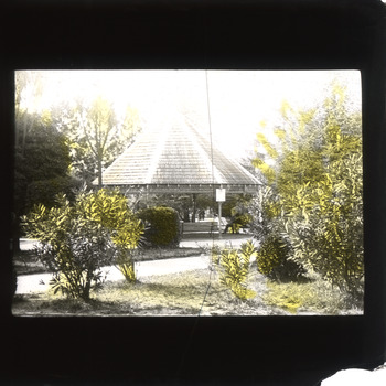 A monochrome photograph within a square frame featuring a rotunda and a man dressed in a suit sitting on a bench in a garden setting.