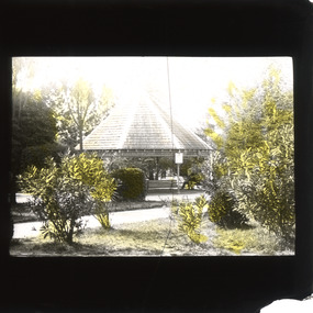 A monochrome photograph within a square frame featuring a rotunda and a man dressed in a suit sitting on a bench in a garden setting.
