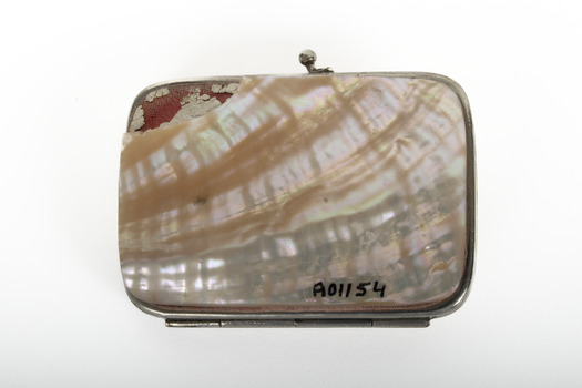 A small rectangular purse with silver edging and half clasp, covered in white and cream coloured shell with a large chip in the top left revealing red material beneath.