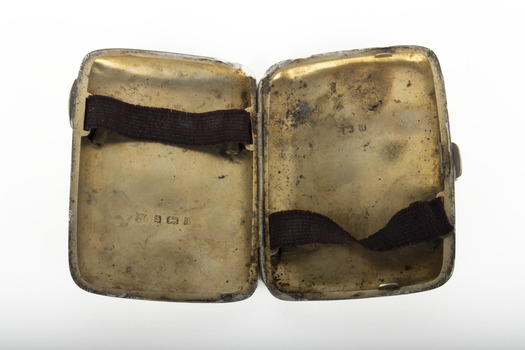 The inside of a silver cigarette case with gold wash, a brown elastic strap on each side and hallmarks stamped on each side.
