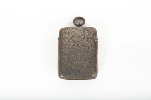 A small silver container featuring an engraved design across it's surface and a ring at the top of the lid.