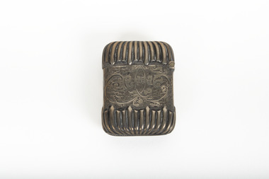 A small silver container featuring an engraved design around the middle surface with a rib design on the top and bottom.