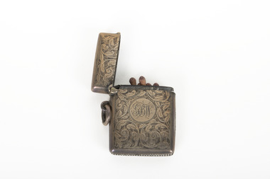 A silver coloured match holder made from engraved steel with detailed scrollwork, it is narrow and fits a single or double row of matches. It features a circular monogram in the centre. The top of the matchbox is hinged to allow for access to the contents. There is a loop at the side to allow for it to be attached to a string or wire or belt.