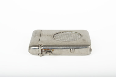 A plain silver coloured matchbook holder made from unornamented metal, it is narrow and fits a single or double row of matches. There is a circular setting on the obverse face with raised crosshatching. It is hinged at the top and has a metal loop for attachment. 