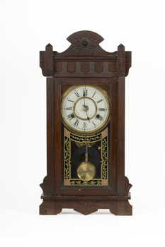 A deep brown wooden striking mantle clock with decorative carvings featuring a glass door with black, gold, and red etched detail. The clock face is white with gold rims and black roman numerals marked.