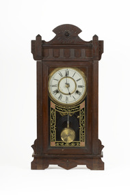 A deep brown wooden striking mantle clock with decorative carvings featuring a glass door with black, gold, and red etched detail. The clock face is white with gold rims and black roman numerals marked.