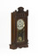 A deep brown wooden striking mantle clock with decorative carvings featuring a glass door with black, gold, and red etched detail. The clock face is white with gold rims and black roman numerals marked.