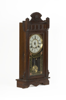 A deep brown wooden striking mantle clock with decorative carvings featuring a glass door with black, gold, and red etched detail. The clock face is white with gold rims and black roman numerals marked.
