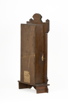 Rear side angle of wooden striking mantle clock showing shepherd's hook closure and an aged maker's label affixed to the back.